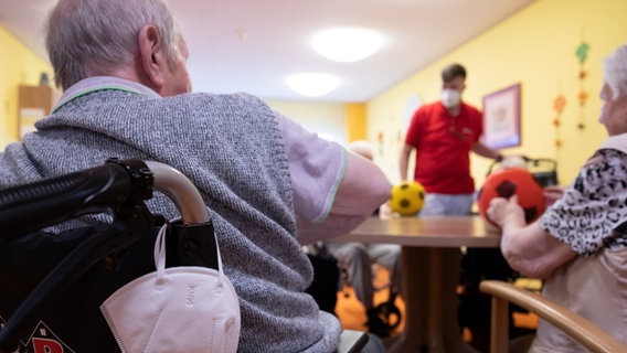 Bewohner spielen in einem Altenheim mit Bällen auf einem Tisch. An einem Rollstuhl hängt eine FFP2-Maske. © dpa picture alliance Foto: Bernd Weißbrod