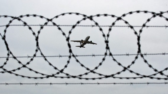 Ein Flugzeug startet, fotografiert durch Stacheldraht am Flughafenzaun. © picture alliance/dpa Foto: Julian Stratenschulte