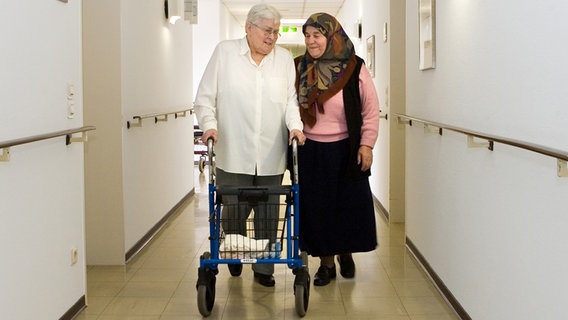 Türkische Frau besucht Seniorin im Altenheim, die einen Rollator schiebt. © picture alliance / Caro | Dobiey Foto: picture alliance / Caro | Dobiey