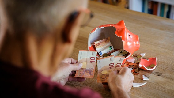 Ein alter Mann sitzt zu Hause am Tisch vor einem zerbrochenen Sparschwein und zählt Geld. © picture alliance Foto: Thomas Trutschel