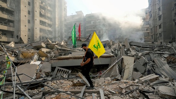 Ein Mann trägt eine Hisbollah-Fahne, während er über die Trümmer seiner zerstörten Wohnung nach einem israelischen Luftangriff auf den Stadtteil Dahieh in Beirut läuft. © AP/dpa Foto: Hassan Ammar