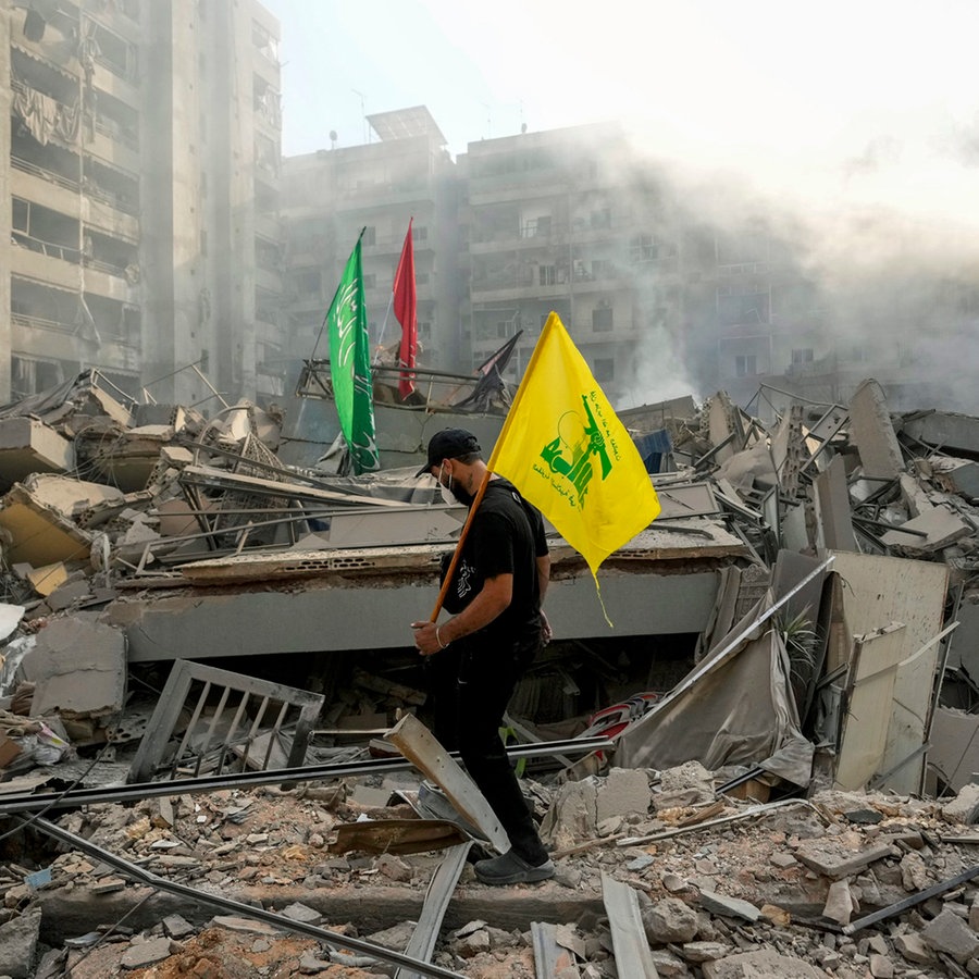 Ein Mann trägt eine Hisbollah-Fahne, während er über die Trümmer seiner zerstörten Wohnung nach einem israelischen Luftangriff auf den Stadtteil Dahieh in Beirut läuft. © AP/dpa Foto: Hassan Ammar