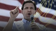 Ron DeSantis, der Gouverneur von Florida, steht vor der amerikanischen Flagge und spricht in ein Mikrofon. Er hält seinen Zeigefinger in die Luft © picture alliance/ dpa/ AP Foto: Andy Barron