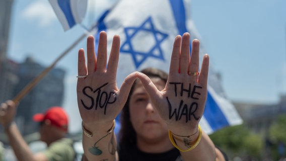 Eine israelische Demonstrantin hält ihre Hände mit der Aufschrift "Stop the War" hoch vor einer israelischen Flagge im Hintergrund, während einer Kundgebung in Tel Aviv. © dpa Foto: Ilia Yefimovich
