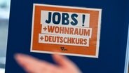 Auf einer Jobmesse für Geflüchtete steht ein Schild mit der Aufschrift Jobs! + Wohnraum + Deutschkurs © picture alliance Foto: Jens Kalaene