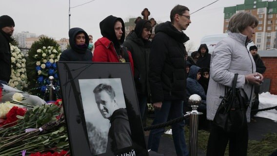 Menschen gehen an einem Porträt des russischen Oppositionellen Alexej Nawalny während der Beerdigungszeremonie auf dem Friedhof Borisowskoje in Moskau vorbei. © Uncredited/AP/dpa 