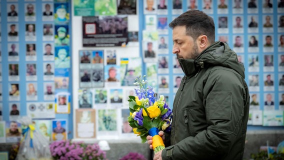 Der ukrainische Präsident Wolodymyr Selenskyj steht für eine Schweigeminute, bevor er Blumen an der Mauer zum Gedenken an die gefallenen Verteidiger der Ukraine auf dem Mykhailivska-Platz niederlegt. © picture alliance / ZUMAPRESS.com | Ukraine Presidency 