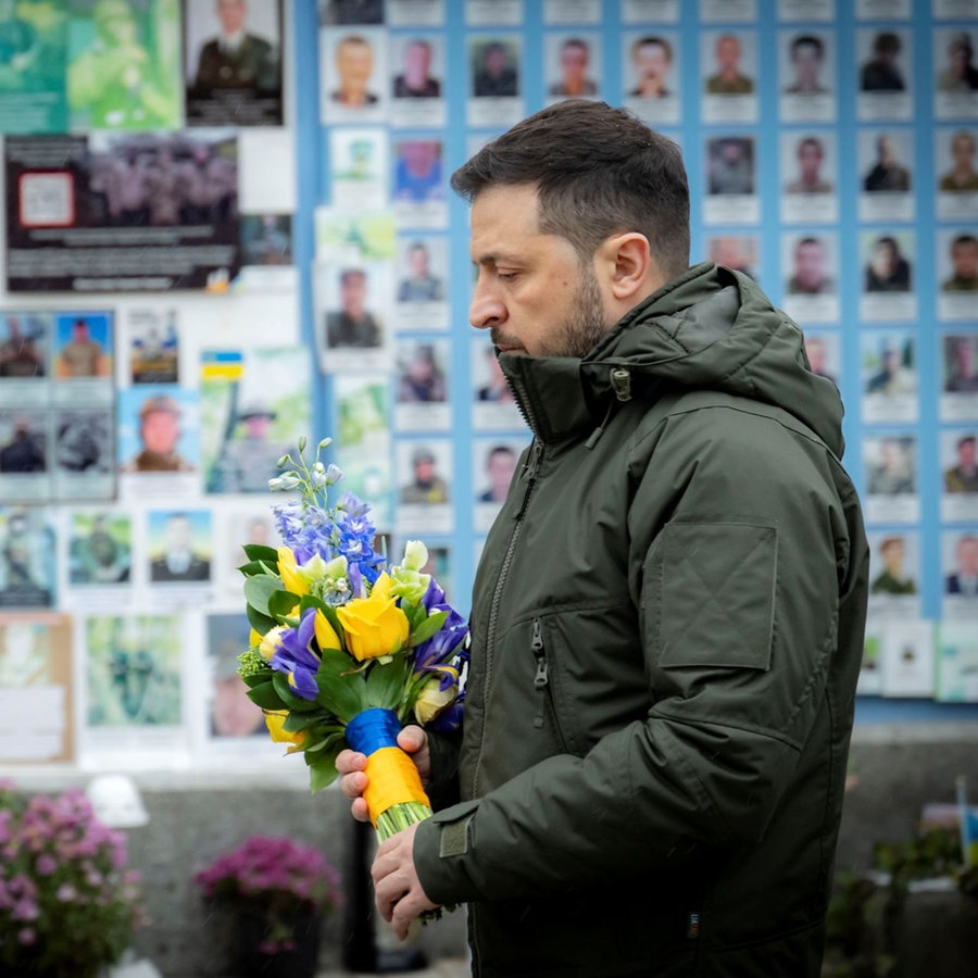 Der ukrainische Präsident Wolodymyr Selenskyj steht für eine Schweigeminute, bevor er Blumen an der Mauer zum Gedenken an die gefallenen Verteidiger der Ukraine auf dem Mykhailivska-Platz niederlegt. © picture alliance / ZUMAPRESS.com | Ukraine Presidency 