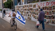 Passanten kommen an Fotos der im Gazastreifen festgehaltenen Geiseln, die an den Wänden des Geiselplatzes inTel Aviv (Israel) angebracht sind, vorbei. © AP/dpa Foto: Oded Balilty