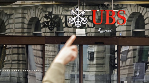 Ein Mann zeigt auf die Logos der Schweizer Banken Credit Suisse und UBS am Paradeplatz in Zürich. © KEYSTONE/dpa Foto: Michael Buholzer