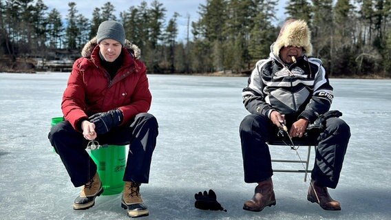 Ingo Zamperoni sitzt in den USA zusammen mit Coach auf einem zugefrorenen See und angelt in einem ins Eis geschlagenen Loch. © NDR Foto: Martin Kobold