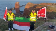 Zwei Post-Mitarbeiter auf Helgoland stehen mit Fahnen der Gewerkschaft ver.di und einer Helgoland-Fahne an der Küste. © Tim Pyschny 