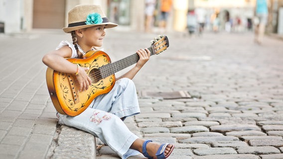 Ein Mädchen mit Hut sitzt auf einer Bordsteinkante und spielt Gitarre © Panther Media 