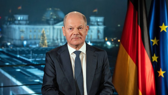 Bundeskanzler Olaf Scholz (SPD) sitzt während eines Fototermins nach der Aufzeichnung seiner Neujahrsansprache im Kanzleramt vor der Kulisse des von Scheinwerfern angestrahlten Reichstages. © Soeren Stache/dpa 