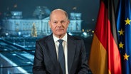 Bundeskanzler Olaf Scholz (SPD) sitzt während eines Fototermins nach der Aufzeichnung seiner Neujahrsansprache im Kanzleramt vor der Kulisse des von Scheinwerfern angestrahlten Reichstages. © Soeren Stache/dpa 