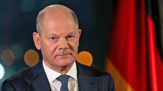 Bundeskanzler Olaf Scholz (SPD) sitzt während eines Fototermins nach der Aufzeichnung seiner Neujahrsansprache im Kanzleramt vor der Kulisse des von Scheinwerfern angestrahlten Reichstages. © Soeren Stache/dpa 