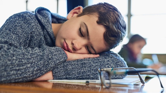 Ein müder Schüler liegt mit Kopf auf einem Schultisch. © IMAGO / YAY Images 