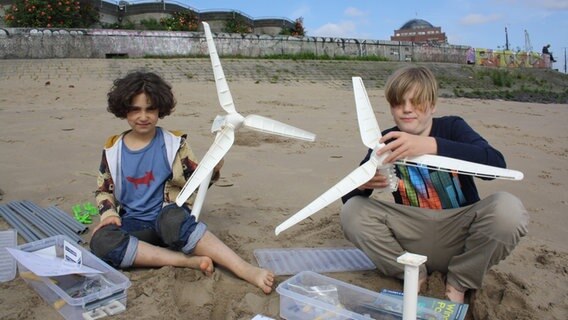 Zwei junge Kinder sitzen am Strand und halten Windräder in den Händen. © NDR Mikado/Sahin 