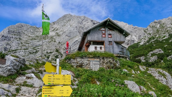 Eine Bergsteigerhütte in den Alpen, davor gelbe Schilder und eine grüne Flagge, dahinter die Alpen © NDR Foto: Jörg Wunram