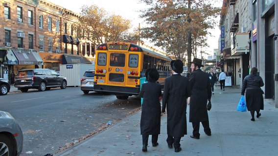 Drei in schwarz gekleidete Männer gehen in den USA auf einem Bürgersteig, daneben ist ein gelber Bus © NDR Foto: Jörn Pissowotzki