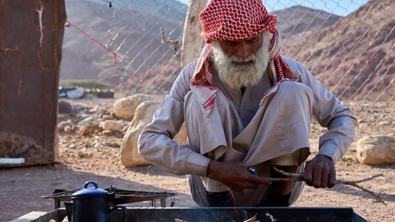 Ein Mann mit rotem Kopftuch und weißem Gewand hockt in der Wüste und zerkleinert Kaffeebohnen © NDR / Florian Guckelsberger Foto: Florian Guckelsberger