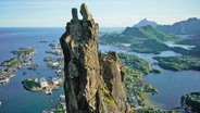 Ein schmaler hoher Felsen ragt über einem Landstück der Lofoten empor, im Hintergrund sind Behausungen und das Meer zu erkennen © NDR Foto: Johannes Koch/Schulze