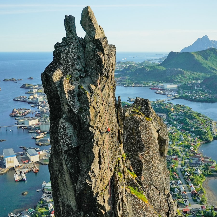 Ein schmaler hoher Felsen ragt über einem Landstück der Lofoten empor, im Hintergrund sind Behausungen und das Meer zu erkennen © NDR Foto: Johannes Koch/Schulze