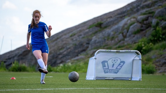 Eine junge Frau spielt Fußball und kickt ein Ball in ein niedriges Tor vor einer Bergkulisse der Lofoten © NDR Foto: Marco Schulze / Johannes Koch