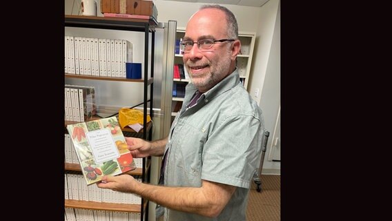 Ein Mann steht seitlich, lächelt und hält ein Buch über Gemüse in der Hand © NDR / Jörn Pissowotzki Foto: Jörn Pissowotzki