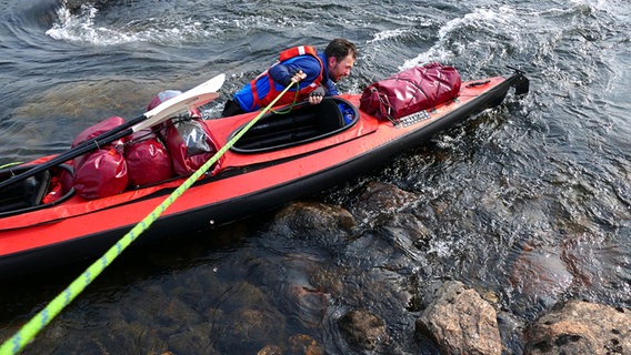 Ein Mann watet durch einen Fluss und kontrolliert ein Faltkajak, das an einer Leine hängt © NDR Foto: Achill Moser und Aaron Moser