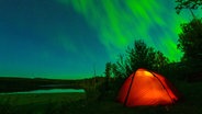 Ein von innen beleuchtetes kleines Zelt zeichnet sich in hellorange gegen den in Polarlichter beleuchteten Nachthimmel ab © NDR Foto: Achill Moser und Aaron Moser