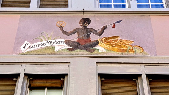 Auf einer Hausfassade in Zürich ist ein lachender Mensch mit dunkler Hautfarbe im Schneidersitz abgebildet - darüber steht "Zum kleinen Mohren" (Schriftzug von 1671) © NDR Foto: Michael Marek