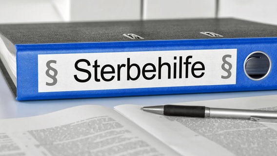 Ein Aktenordner mit der Aufschrift "Sterbehilfe" liegt auf einem Schreibtisch, im Vordergrund Formulare und ein Kugelschreiber. (Symbolbild Sterbehilfe) © picture alliance / Shotshop | Zerbor 