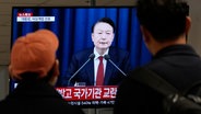 Menschen schauen an einem Busbahnhof auf einen Fernsehbildschirm, der die im Fernsehen übertragene Ansprache des südkoreanischen Präsidenten Yoon Suk Yeol zeigt. © Ahn Young-joon/AP/dpa 