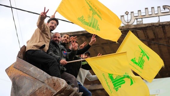 Männer, die in der Schaufel eines Bulldozers sitzen, schwenken Hisbollah-Fahnen, während sie die Waffenruhe zwischen Israel und der Hisbollah feiern, die am Mittwoch (27.11.2024) begonnen hat. © dpa/AP Foto: Mohammed Zaatari