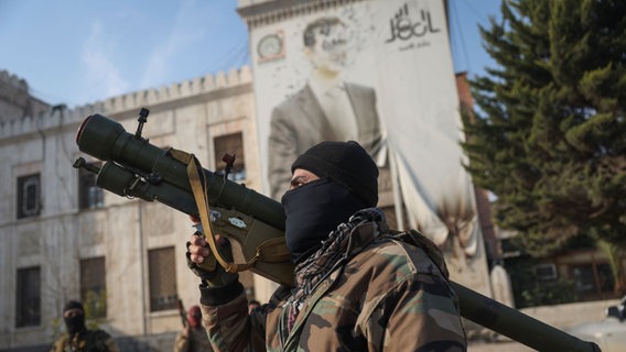 Ein syrischer Oppositionskämpfer hält einen Raketenwerfer vor dem Büro der Provinzregierung, an dessen Fassade ein Bild des syrischen Präsidenten Baschar Assad von Kugeln durchlöchert ist, nach der Übernahme der Stadt. © picture alliance/dpa/AP | Ghaith Alsayed Foto: Ghaith Alsayed