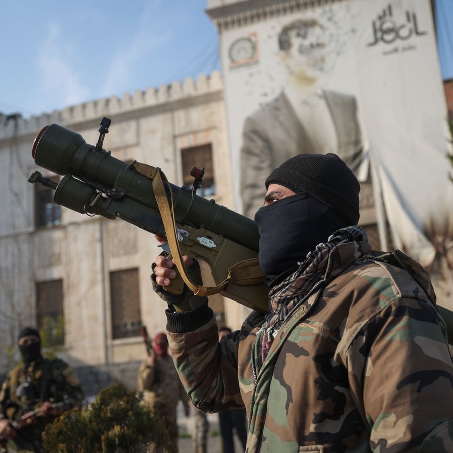 Ein syrischer Oppositionskämpfer hält einen Raketenwerfer vor dem Büro der Provinzregierung, an dessen Fassade ein Bild des syrischen Präsidenten Baschar Assad von Kugeln durchlöchert ist, nach der Übernahme der Stadt. © picture alliance/dpa/AP | Ghaith Alsayed Foto: Ghaith Alsayed