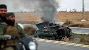 Syrische Oppositionskämpfer fahren an einem brennenden gepanzerten Fahrzeug der Regierung südlich von Hama, Syrien, vorbei. © dpa-Bildfunk/AP Foto: Ghaith Alsayed