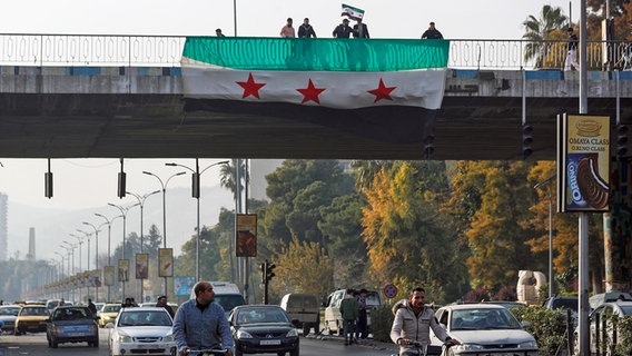 Syrer zeigen an einer Brücke die Fahne der syrischen Opposition, während sie in Damaskus feiern. © picture alliance/dpa/AP Foto: Omar Sanadiki