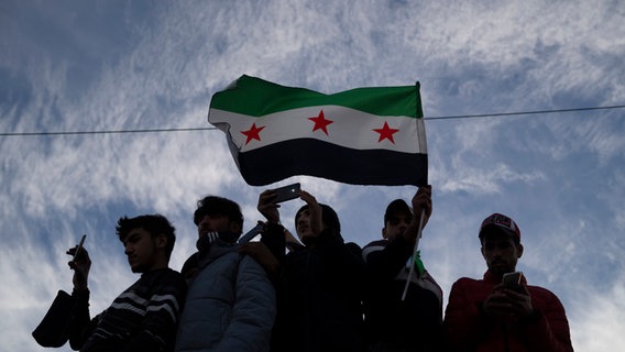 Unter einer syrischen Flagge versammeln sich mehrere Menschen. © dpa Bildfunk Foto: Leo Correa