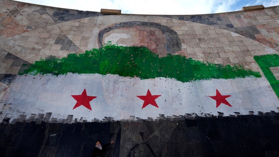 Ein syrischer Junge steht unter einem riesigen Porträt des verstorbenen syrischen Präsidenten Al-Assad, übermalt mit den Farben der sogenannten "revolutionären" Flagge, in Damaskus. © Leo Correa/AP/dpa 