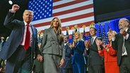 Donald Trump reckt seine Faust in die Höhe und geht mit seiner Frau Melania auf einer Bühne an klatschenden Unterstützern vorbei. © dpa bildfunk/AP Foto: Evan Vucci