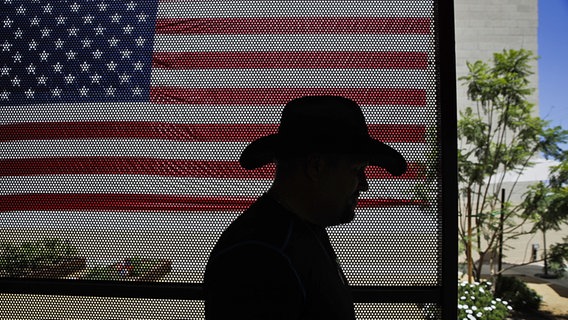 Vor einer amerikanischen Flagge ist die Silhouette eines Mannes zu sehen, der einen Cowboy-Hut trägt. © dpa picture alliance Foto: Jae C. Hong