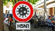 Vor einem Café sitzen Menschen, davor steht ein Warnschild mit einem Virussymbol und der Aufschrift "H5N1". (Symbolbild) © picture alliance / CHROMORANGE | Michael Bihlmayer 