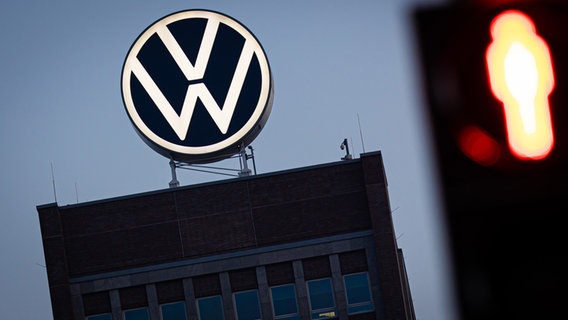 Das VW-Logo leuchtet auf dem Dach des Markenhochhauses im Volkswagen Stammwerk am frühen Morgen hinter einer rot zeigenden Fußgänger-Ampel. © picture alliance Foto:  Moritz Frankenberg