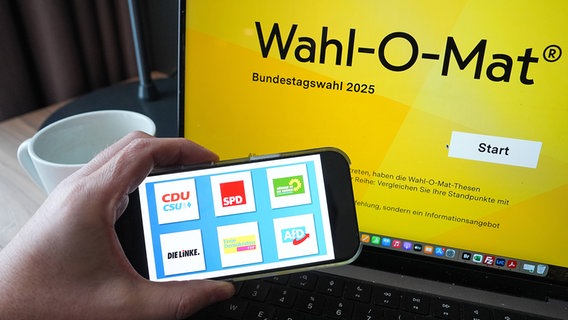 Auf einem Smartphone sind verschiedene Partei-Logos zu sehen, dahinter auf einem Laptop die Startseite des Wahl-O-Mat. © IMAGO Images/Political-Moments 