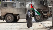 Ein Mann steht mit einer palästinensischen Flagge in der Hand vor einem israelischen Militärfahrzeug im besetzten Westjordanland. ©  Nasser Ishtayeh/SOPA Images via ZUMA Press Wire/dpa 