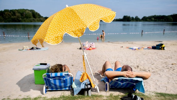 29.08.2024, Niedersachsen, Oldenburg: Ein Paar sitzt bei sonnigem Wetter und hohen Temperaturen auf zwei Liegen unter einem Sonnenschirm im Sand am Kleinen Bornhorster See. © dpa Foto: Hauke-Christian Dittrich