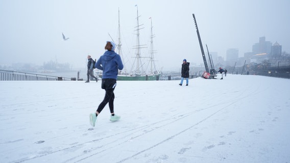 Eine Frau joggt in Hamburg auf der schneebedeckten Jan-Fedder-Promenade am Hafen. © Marcus Brandt/dpa Foto: Marcus Brandt