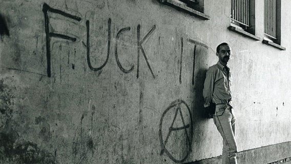 Ein Mann lehnt an einer Hauswand, auf der groß "Fuck it" geschrieben steht. © NDR 